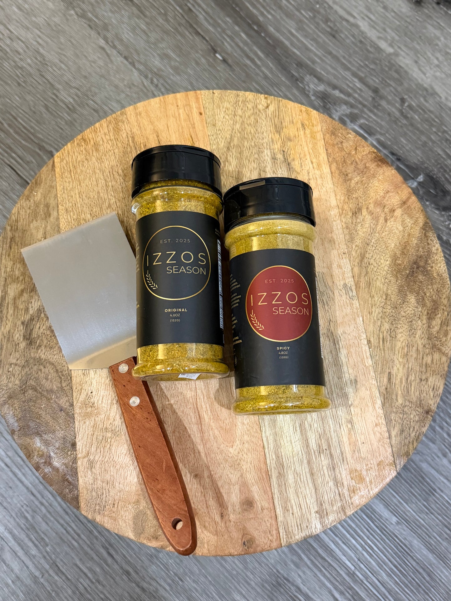 Izzo's Season Original and Spicy all-purpose seasoning bottles with a wooden grill scraper thats stainless steel